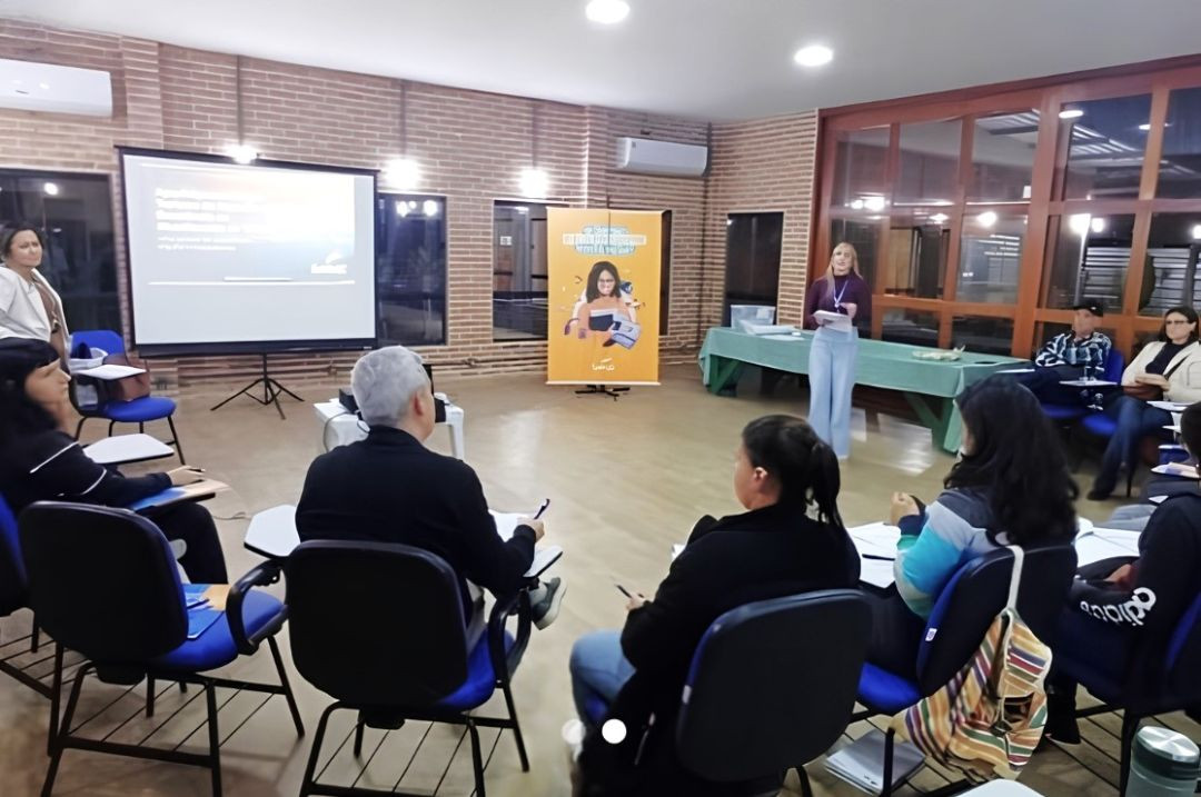 Foto horizontal em ângulo aberto mostrando uma sala de aula ou auditório com paredes de tijolos e cadeiras universitárias azuis dispostas em semicírculo. No fundo, à direita, uma palestrante loira, vestindo blusa roxa e calça clara, fala de pé perto de um banner amarelo. À esquerda, há um projetor exibindo slides em uma tela branca. Participantes sentados prestam atenção à apresentação.