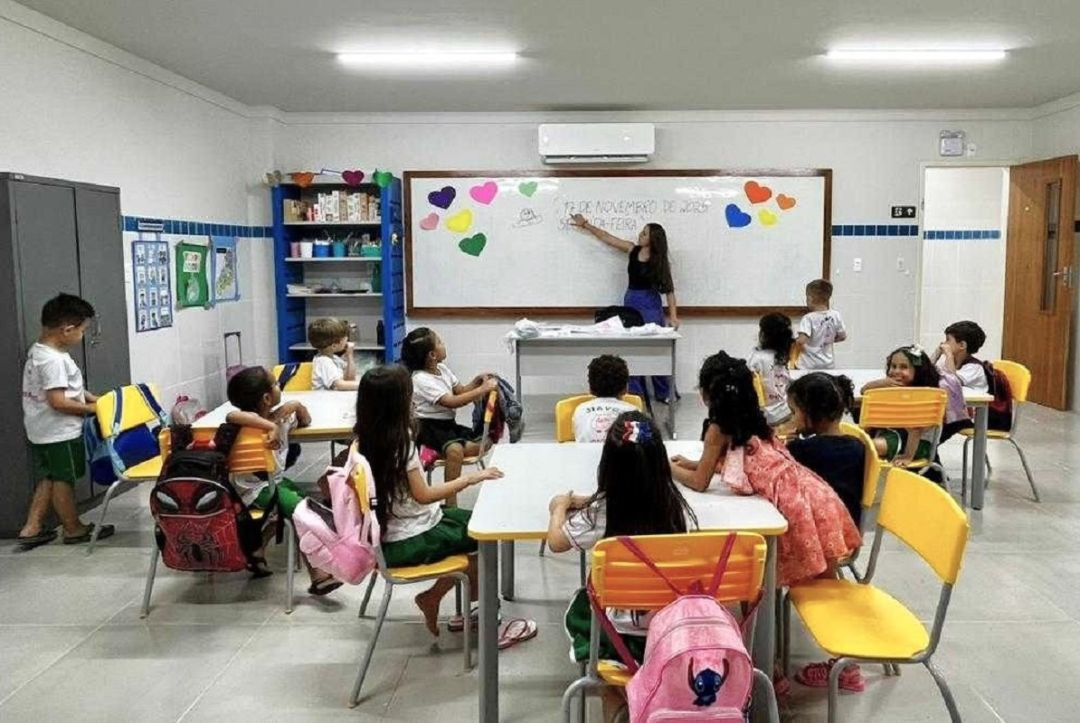 Sala de aula de uma escola municipal de educação infantil com paredes brancas e detalhes em azulejos azuis. Ao fundo, uma professora em pé aponta para uma lousa branca decorada com corações coloridos de papel, onde se lê 