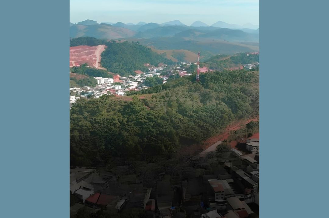Vista aérea panorâmica de uma área urbana e montanhosa em Iúna. Em primeiro plano, no quadrante inferior, há um aglomerado de casas. Logo acima, destaca-se uma extensa colina coberta por vegetação densa e árvores verdes, onde está instalada uma torre de transmissão vertical nas cores vermelha e branca. Ao fundo, a paisagem exibe relevos montanhosos sob um céu claro, evidenciando o contraste entre o crescimento urbano e a área verde preservada.