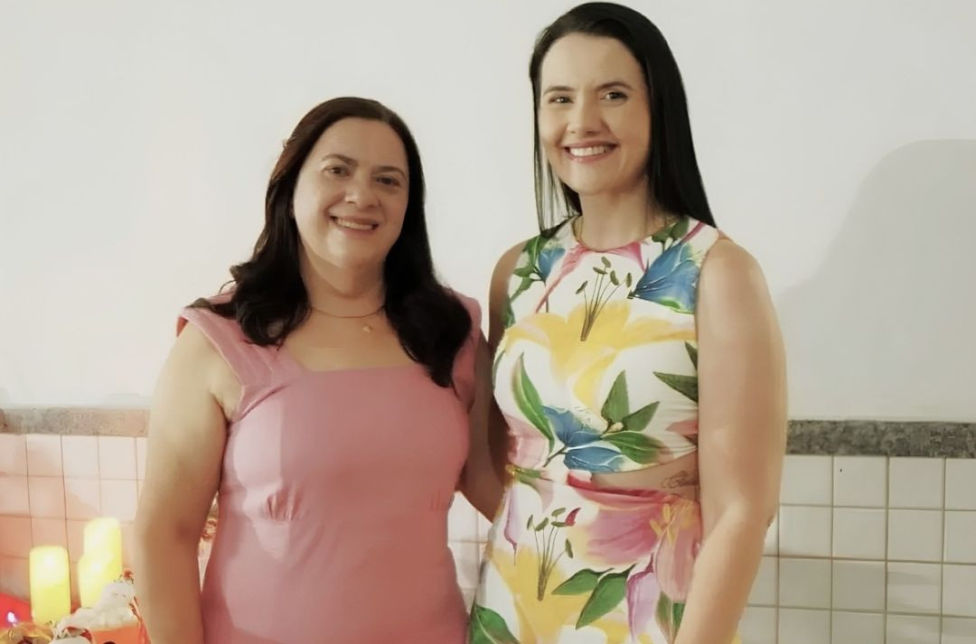 Fotografia em plano médio de duas mulheres sorrindo, posicionadas lado a lado em um ambiente interno. À esquerda, uma mulher de cabelos escuros e compridos veste um vestido liso na cor rosa claro. À direita, outra mulher, também de cabelos escuros e compridos, veste um vestido sem mangas com estampa floral colorida em tons de amarelo, rosa, verde e azul. Ao fundo, vê-se uma parede branca, uma faixa de revestimento cerâmico claro e, no canto inferior esquerdo, parte de velas acesas.