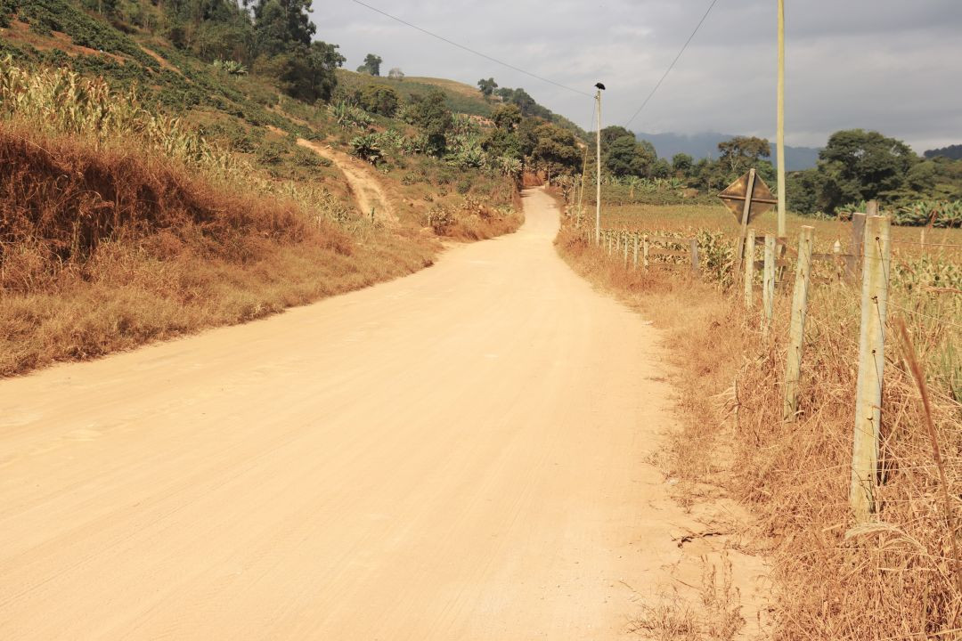 Vista frontal de uma estrada de terra batida, larga e de coloração clara e seca. À esquerda, há um barranco com vegetação rasteira e pasto seco. À direita, uma cerca de arame farpado com mourões de madeira margeia uma plantação de milho. Ao fundo, veem-se morros com plantações e vegetação nativa sob um céu parcialmente nublado.