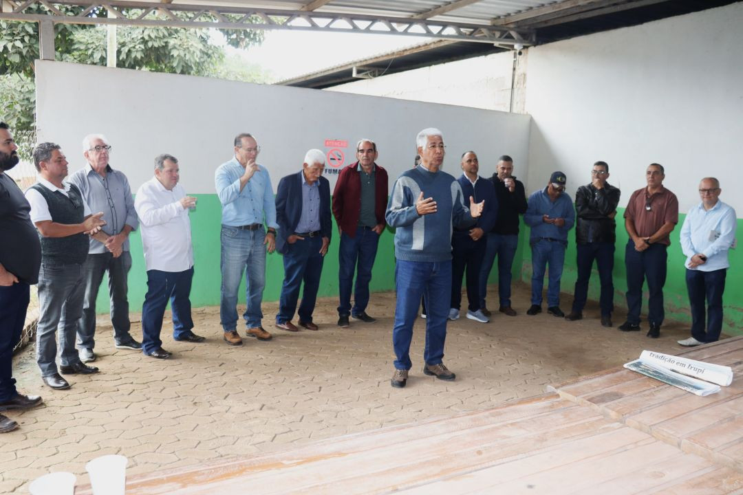 Vista lateral de um homem de cabelo grisalho e suéter azul escuro gesticulando enquanto fala aos presentes. Ele está posicionado no centro de um galpão com piso de blocos de concreto sextavados. À esquerda, o secretário Enio Bergoli e outras autoridades observam o discurso.