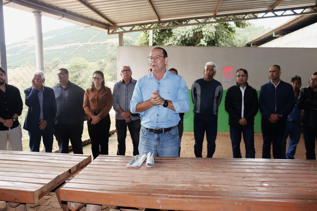 Homem de óculos, camisa social azul clara e calça jeans fala ao público com as mãos juntas, sob uma estrutura de galpão coberto com telhas de zinco. Atrás dele, um grupo de homens e uma mulher acompanham o pronunciamento. Sobre uma mesa de madeira em primeiro plano, há jornais dobrados.