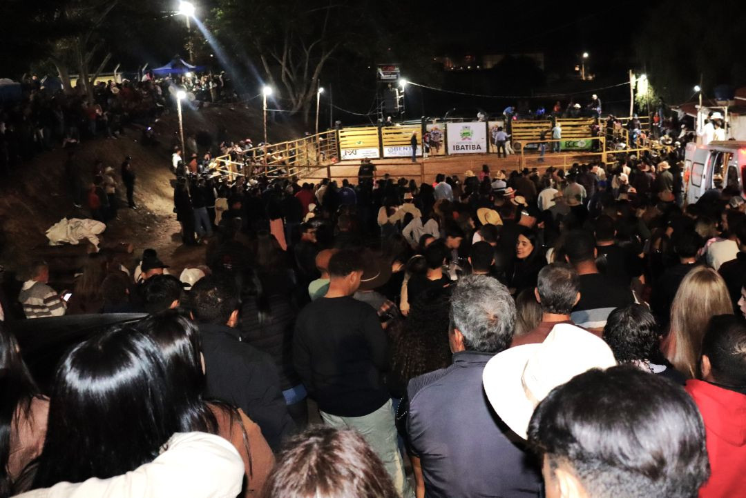 Imagem em plano geral noturno mostrando a arena do rodeio da Festa da Associação Carangolas. A arena está cercada por uma cerca e preenchida por uma multidão de espectadores assistindo à competição de rodeio no centro. Há iluminação no local e a placa 