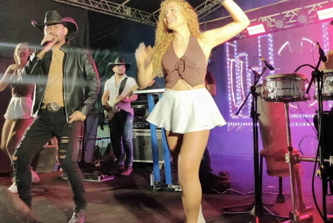 Foto capturada durante uma apresentação musical da 5ª Festa da Associação Carangolas. No centro, uma jovem com cabelos cacheados e roupas claras dança no palco, com iluminação cênica. Ao fundo, músicos tocam instrumentos, incluindo um percussionista e um guitarrista, sob uma estrutura de treliças de metal.