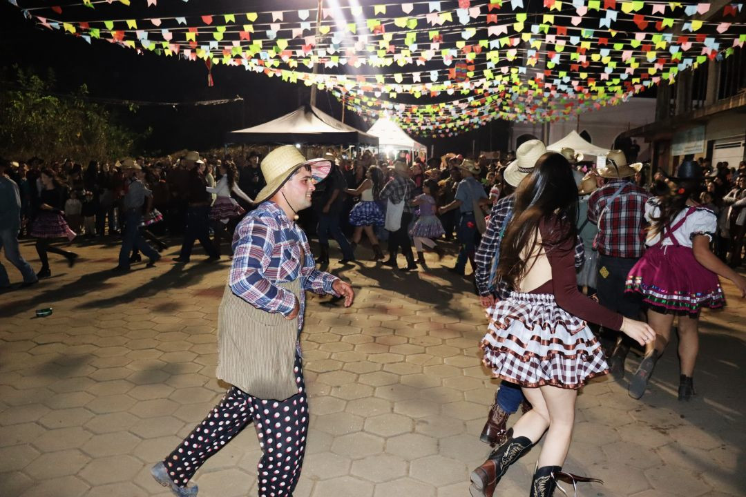 Foto capturada durante a apresentação de quadrilha na 5ª Festa da Associação Carangolas. Dançarinos vestidos com trajes típicos juninos e chapéus de palha dançam em um espaço pavimentado sob um teto de bandeirinhas coloridas e luzes. O casal principal da dança está em destaque no centro da imagem.