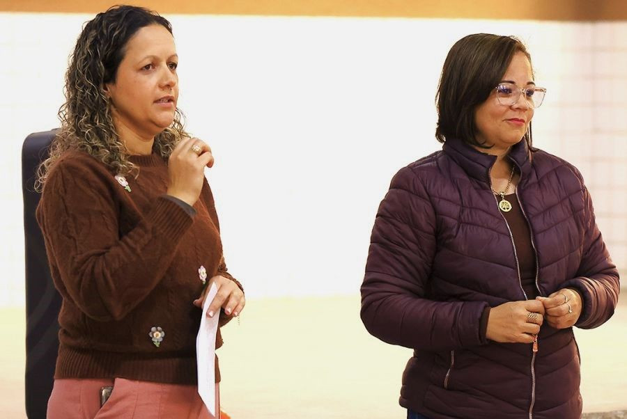 Close-up das duas palestrantes (psicóloga e assistente social) do CREAS. Elas estão em pé, segurando papéis e microfones, falando com atenção para os participantes que não aparecem na imagem. Ambas vestem roupas adequadas para o evento.