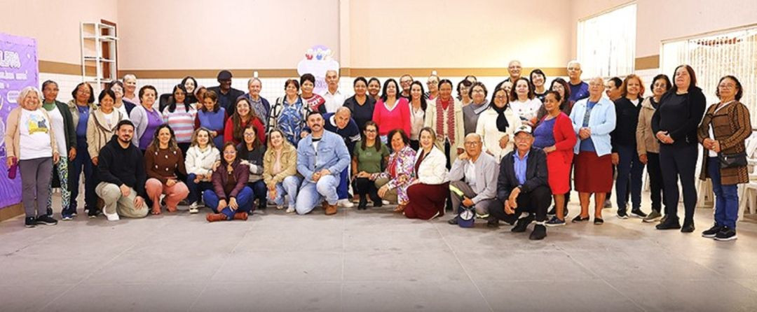 Foto panorâmica capturando todos os participantes e organizadores da ação do Junho Violeta. O grupo é majoritariamente formado por idosos, posando em frente ao palco onde uma faixa da campanha está visível.