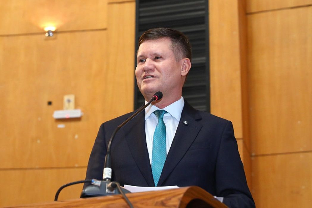 Homem de cabelos castanhos curtos, vestindo terno azul-marinho, camisa clara e gravata verde-esmeralda com texturas. Ele está em pé atrás de um púlpito de madeira com microfone fixo, discursando voltado para a esquerda. Ao fundo, painéis de madeira cobrem a parede do plenário.