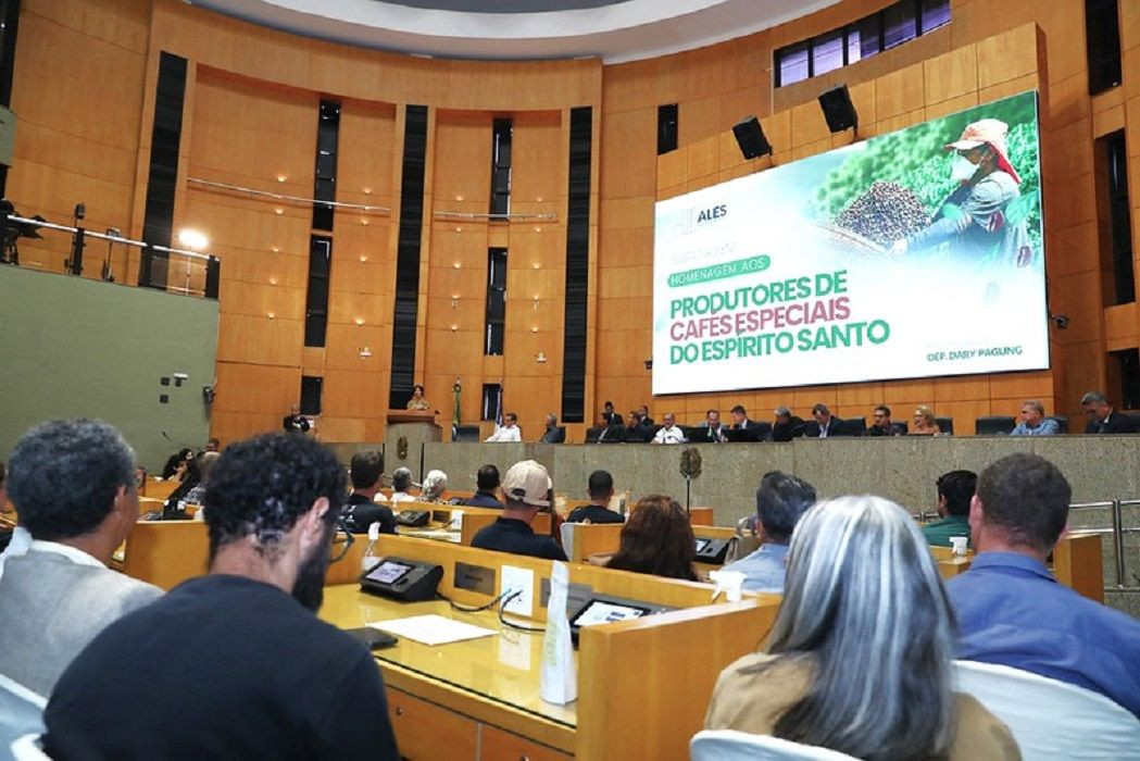 Vista interna do plenário da Assembleia Legislativa a partir da perspectiva do público. No fundo, uma grande bancada com autoridades sentadas e um imenso telão digital exibe a frase 