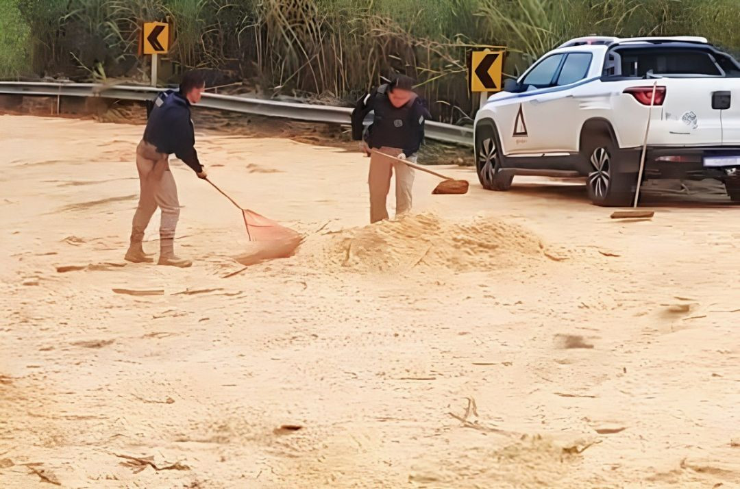 Fotografia em plano médio mostrando dois agentes da Polícia Rodoviária Federal (PRF) trabalhando na limpeza do asfalto da BR-262. Eles vestem uniformes escuros e calças caqui. O agente à esquerda utiliza um rastelo de metal e o da direita utiliza uma enxada para espalhar uma grande quantidade de pó de serra de cor bege claro, que cobre quase toda a pista para conter o vazamento de óleo. Ao fundo, no acostamento à direita, está estacionada uma picape Fiat Toro branca, de costas, com a tampa da caçamba aberta. Há placas de sinalização de curva acentuada à esquerda (setas pretas em fundo amarelo) e uma vegetação alta e verde (capim e árvores) que se estende por um barranco ao fundo, sob a base de um morro.