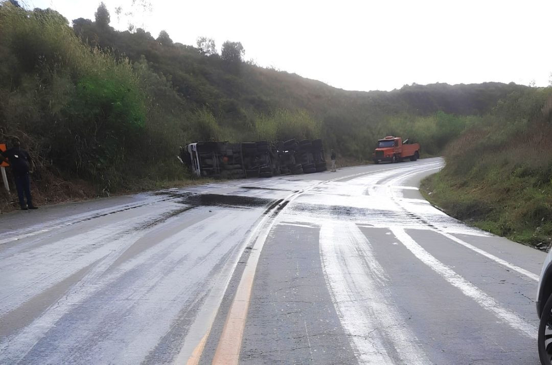 Fotografia em plano aberto da rodovia BR-262 em Ibatiba, mostrando marcas escuras de frenagem e poças de óleo de xisto espalhadas pelo asfalto cinza, que está parcialmente coberto por pó de serra claro. Ao fundo, em uma curva, um caminhão-tanque aparece tombado lateralmente no acostamento, próximo à vegetação e a um guincho laranja. Na parte inferior direita, há uma marca d'água com a data de 30 de junho de 2026.