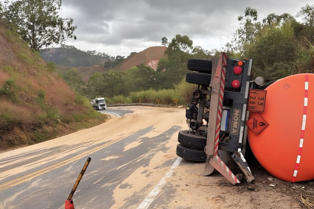 Fotografia em plano médio focada na traseira de um caminhão-tanque laranja da marca Noma, tombado de lado na margem de uma estrada sinuosa. É possível ver os pneus suspensos, a placa do veículo e os painéis de sinalização de carga perigosa com os códigos 33 e 1288 (líquido inflamável). O asfalto da curva está coberto por uma camada de pó de serra marrom claro para absorção do óleo. Ao fundo, a rodovia continua entre montanhas com vegetação rasteira e árvores sob um céu nublado.