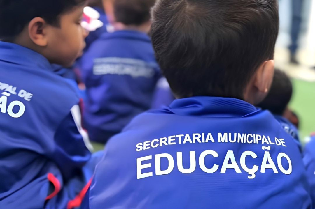 Foto tirada pelas costas de um aluno sentado no chão da escola. Foco nítido na parte traseira da jaqueta do uniforme azul, onde se lê a inscrição em letras brancas de tamanho médio: 