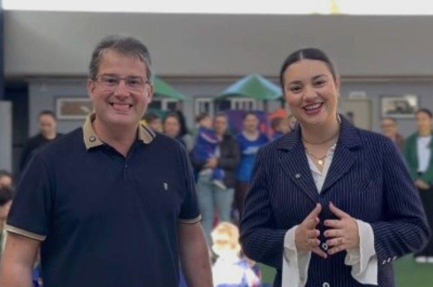 O prefeito Thiaguinho à esquerda, de óculos e camisa polo azul-escura com detalhes em bege na gola. A secretária Esther Simões à direita, com cabelo preso, sorrindo, vestindo blazer azul-escuro risca-de-giz sobre camisa branca e colar dourado, com as mãos articuladas à frente. Ao fundo, de forma desfocada, há uma área externa com brinquedos de playground e pessoas conversando.