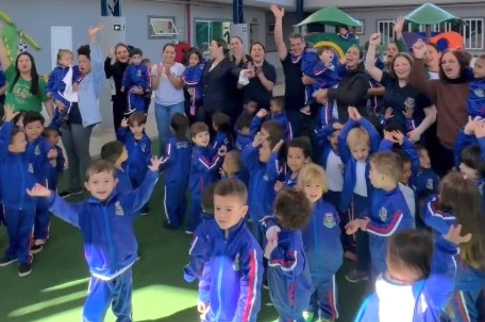 Grupo numeroso de crianças pequenas, alunas da creche municipal, reunidas em um pátio escolar de piso verde. A maioria veste o novo uniforme oficial: um agasalho azul-escuro com faixas vermelhas e brancas nas mangas e o brasão da prefeitura no peito. Algumas crianças e monitoras ao fundo aparecem sorrindo, acenando e aplaudindo em clima de comemoração.