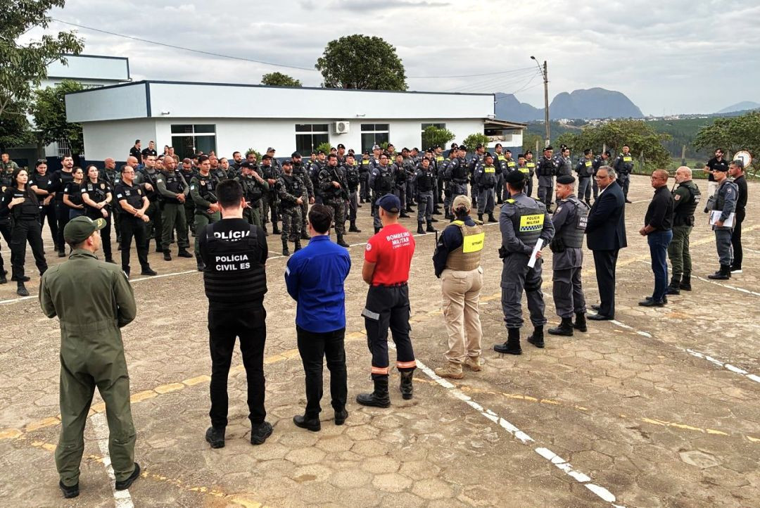 Imagem mostra um grupamento de policiais militares e civis do Espírito Santo, formados em linha na área externa, sob um céu nublado, com prédios ao fundo e uma paisagem montanhosa ao longe. Diversos oficiais estão de uniforme e alguns civis de camisa azul.