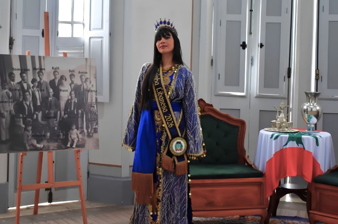 Fotografia em plano médio da Rainha Libanesa 2026, Piscila Simão, com trajes tradicionais libaneses em tons de azul e dourado, usando uma coroa azul e uma faixa transversal de miss onde se lê 
