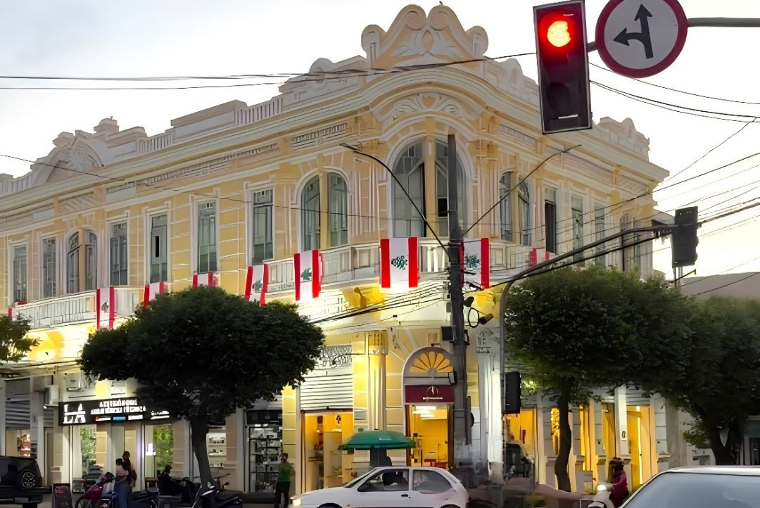 Fotografia de ângulo externo mostrando a fachada do Solar Miguel Simão, prédio histórico de dois andares em estilo eclético, pintado em tons de amarelo claro e branco, localizado em uma esquina de Alegre. Várias bandeiras do Líbano estão estendidas ao longo do guarda-corpo da sacada do segundo andar. Na rua, em frente ao prédio iluminado, há árvores podadas em formato arredondado, um semáforo com a luz vermelha acessa e um carro branco estacionado.
