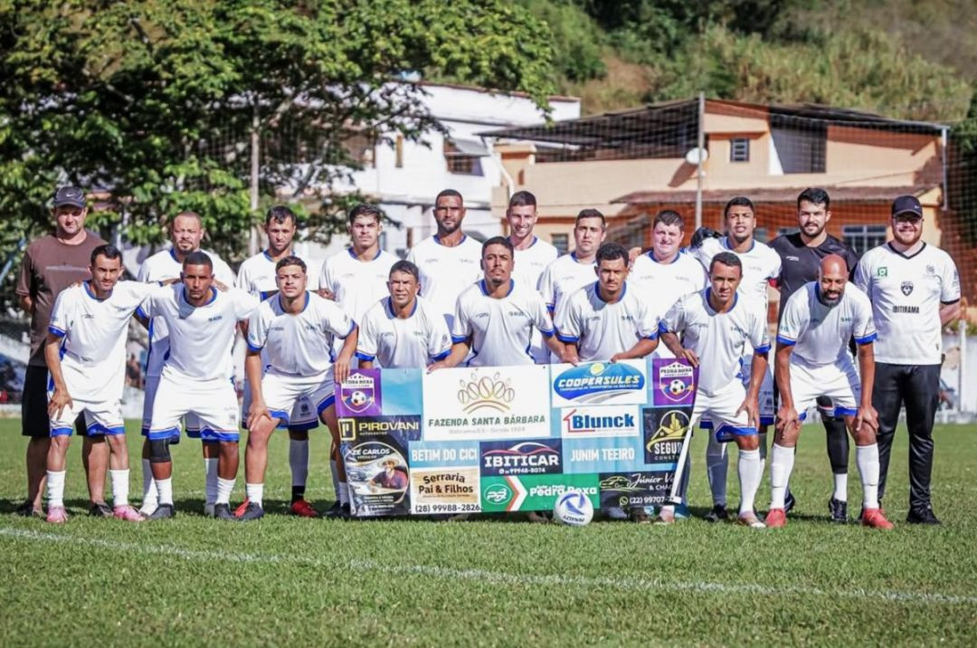 Foto posada da equipe do Pedra Roxa em um campo de futebol gramado. Os jogadores vestem uniforme majoritariamente branco com detalhes em azul e seguram uma faixa horizontal com logotipos de patrocinadores locais. Ao fundo, árvores e casas.