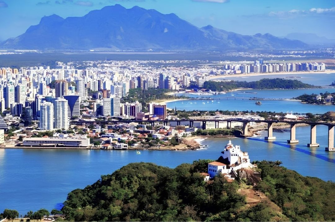 Vista aérea panorâmica da cidade de Vitória, capital do Espírito Santo, exibindo a baía de Vitória com águas azuis, a Terceira Ponte ao fundo conectando à Vila Velha, edifícios urbanos ao longo da orla e o Convento da Penha situado no topo de um morro arborizado em primeiro plano sob céu aberto.