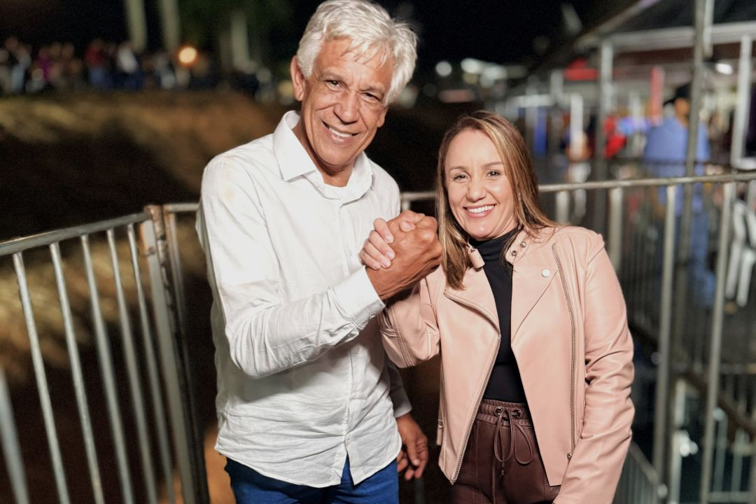 Foto colorida em plano médio de dois adultos sorrindo e olhando para a câmera, com um aperto de mão firme e frontal no centro. À esquerda, o prefeito Dito Silva, um homem mais velho com cabelos brancos, veste camisa branca aberta. À direita, a ex-secretária Emanuela Pedroso, uma mulher com cabelos castanhos, veste jaqueta de couro rosa pálido sobre roupa escura. Ambos estão sorrindo abertamente e apertando a mão um do outro com as duas mãos. O fundo está desfocado, mostrando luzes e estruturas de metal de uma festa noturna, com silhuetas de uma multidão à distância.