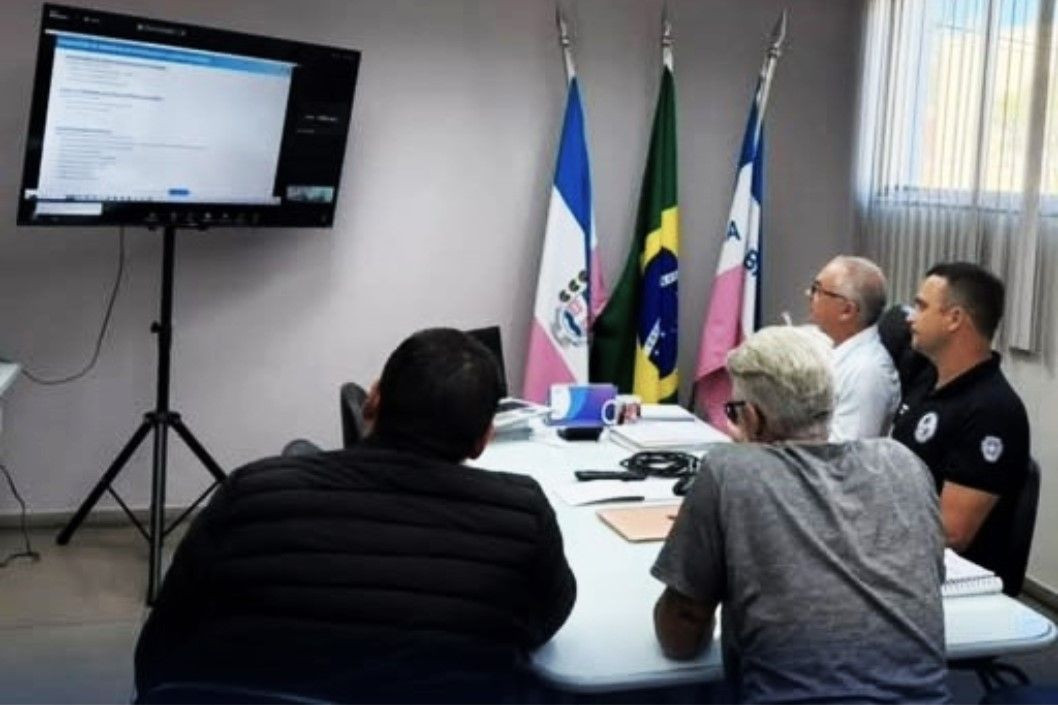 Quatro homens participam de uma reunião em uma sala de escritório. Três deles estão sentados ao redor de uma mesa branca e um está de costas, em primeiro plano. Eles olham em direção a uma tela de televisão fixada em um pedestal à esquerda, que exibe um documento com gráficos e textos. Ao fundo, no canto direito da sala, estão posicionadas três bandeiras oficiais em pedestais: a do município, a do Brasil e a do estado do Espírito Santo.