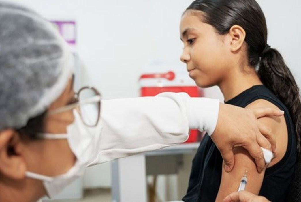 Fotografia em plano médio focada no perfil de uma jovem de cabelos escuros amarrados em um rabo de cavalo, vestindo blusa preta, recebendo uma vacina no braço direito. No primeiro plano, à esquerda e de costas parciais, uma profissional de saúde usando óculos de grau, máscara descartável branca e touca cirúrgica na cabeça segura uma seringa e aplica o imunizante. O fundo da sala de atendimento é claro e exibe de forma desfocada uma caixa térmica vermelha sobre um balcão.