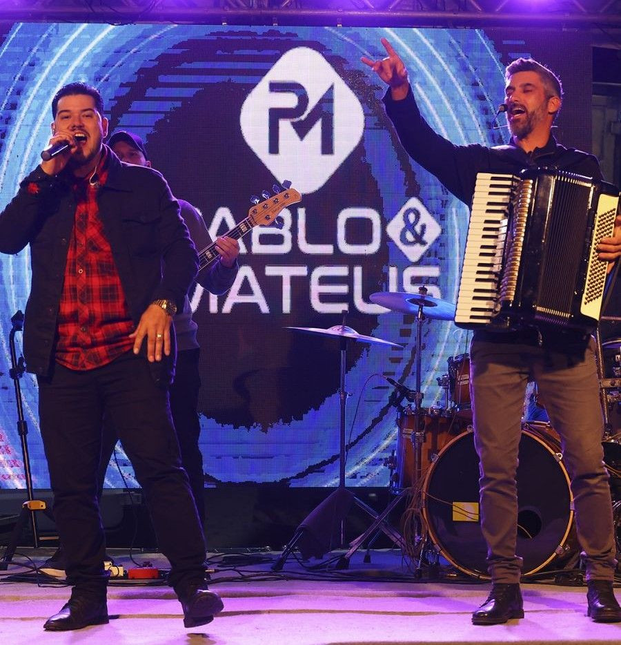 Foto noturna mostrando a dupla sertaneja Pablo e Mateus cantando no palco montado para o encerramento da programação em Pequiá. Um canta e o outro toca sanfona e violão. O fundo do palco tem um painel de LED azul.