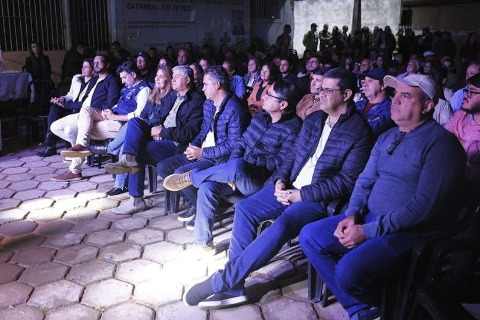 Foto noturna mostrando a plateia da cerimônia de inauguração da UBS em Pequiá. Diversas autoridades e moradores estão sentados em cadeiras de plástico pretas, em uma rua pavimentada, assistindo à solenidade.