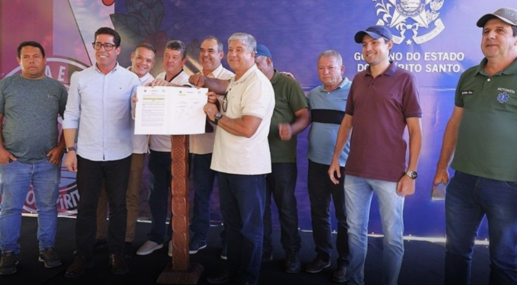Foto de dia durante a assinatura da ordem de serviço para o Campo Bom de Bola. O prefeito Romário Vieira, o deputado Marcelo Santos, e o secretário Weverton Feitosa, junto com outros vereadores e secretários, seguram o documento da ordem de serviço assinada, de pé em frente a um painel azul do Governo do Estado do Espírito Santo.