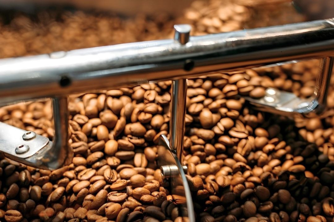 Close-up mostrando o mecanismo de um braço agitador de aço inoxidável operando dentro de um tambor de refrigeração industrial, movendo grãos de café recém-torrados de cor marrom rica.