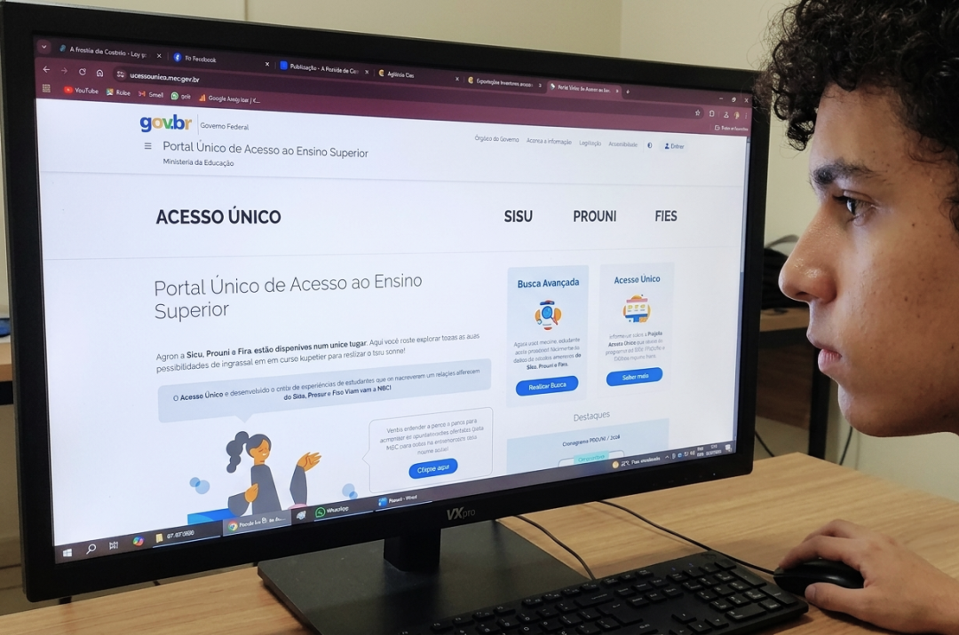 Homem jovem de perfil com cabelos cacheados observa atentamente um monitor de computador. Na tela, está exibida a página inicial do Portal Único de Acesso ao Ensino Superior, com o logotipo do Governo Federal e a seção do Prouni detalhando o cronograma do programa com caixas de texto nas cores azul e cinza sobre fundo branco. O jovem apoia a mão direita no mouse.