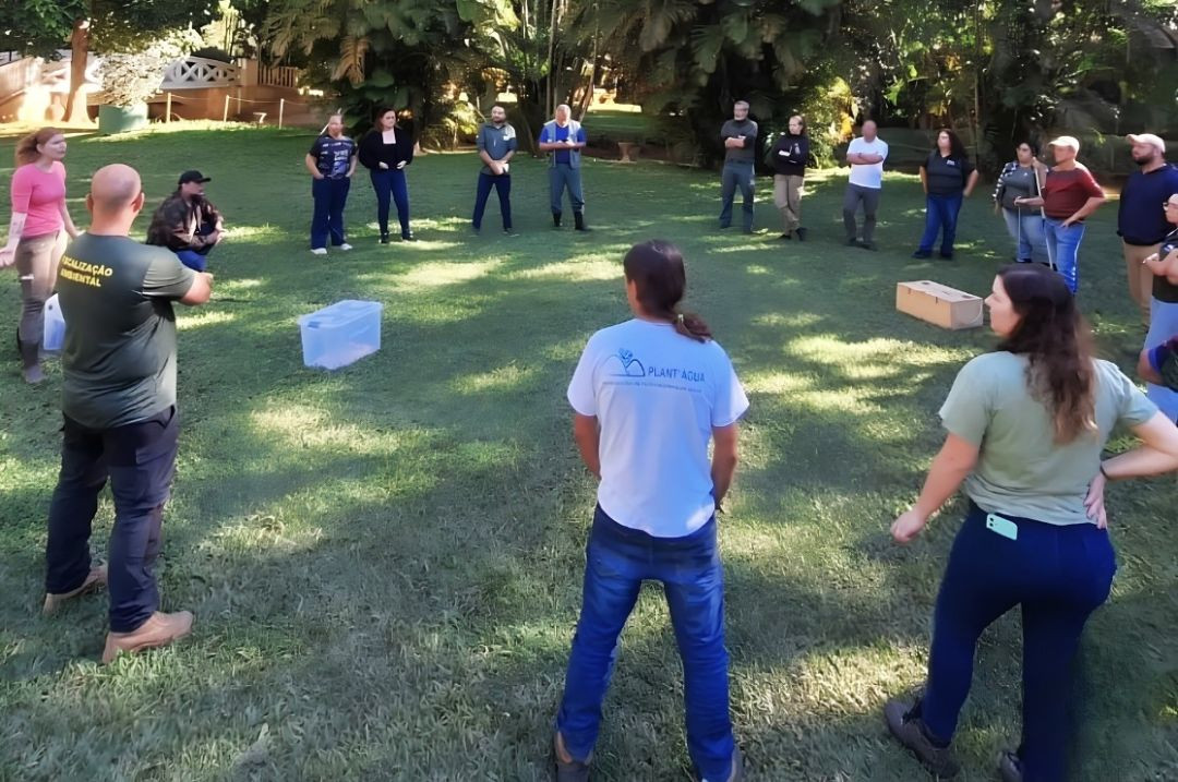 Grupo de cerca de vinte participantes do curso reunidos em círculo em uma área de gramado ensolarada, cercada por árvores. À esquerda, um instrutor vestindo camisa verde com a inscrição 