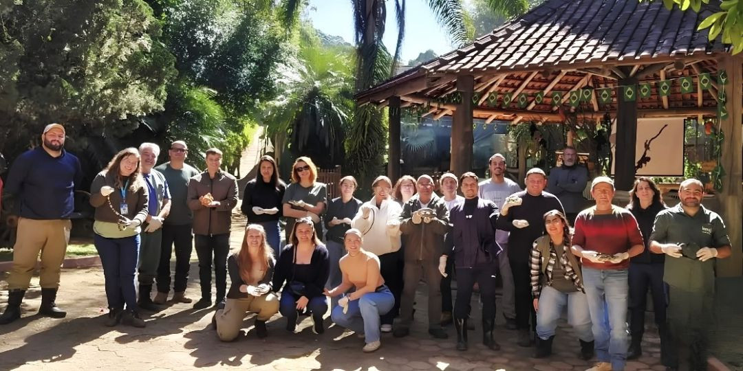 Foto oficial de encerramento com mais de vinte participantes posicionados em frente ao quiosque do Zoopark da Montanha. Alguns utilizam luvas de proteção e seguram pequenos animais ou répteis sob supervisão para registrar o término da capacitação técnica.