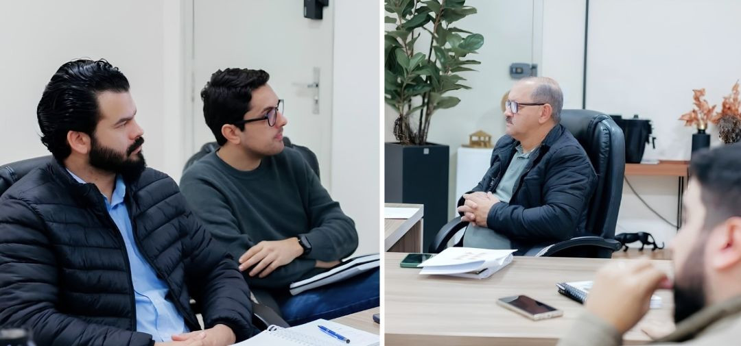 Registro dividido da mesa de reuniões. No lado esquerdo, dois participantes de perfil ouvem atentamente os comunicados, trajando vestimentas de inverno devido ao clima ameno. Do lado direito, um gestor sênior de óculos, bigode e jaqueta preta acolchoada acompanha o debate com as mãos unidas sobre a mesa técnica, ambientado em uma sala administrativa iluminada com porta de vidro ao fundo.