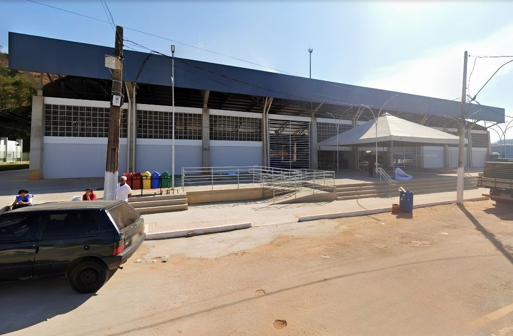 Fachada externa do Centro de Eventos de Guaçuí/ES sob luz solar forte. O edifício é uma estrutura ampla de metal e concreto, com telhado e marquise superior em cinza escuro. Paredes brancas em cobogós (tijolos vazados) compõem a maior parte da frente, seguidas por grandes portões de enrolar em aço. À direita, um gazebo piramidal branco está montado na área de acesso. Há rampas de acessibilidade em concreto com corrimãos metálicos prateados subindo para a entrada. Em primeiro plano, no asfalto em frente à estrutura, vê-se a traseira de um carro verde escuro estacionado e algumas pessoas em pé e sentadas na calçada, além de lixeiras coloridas e postes de luz com fiação.