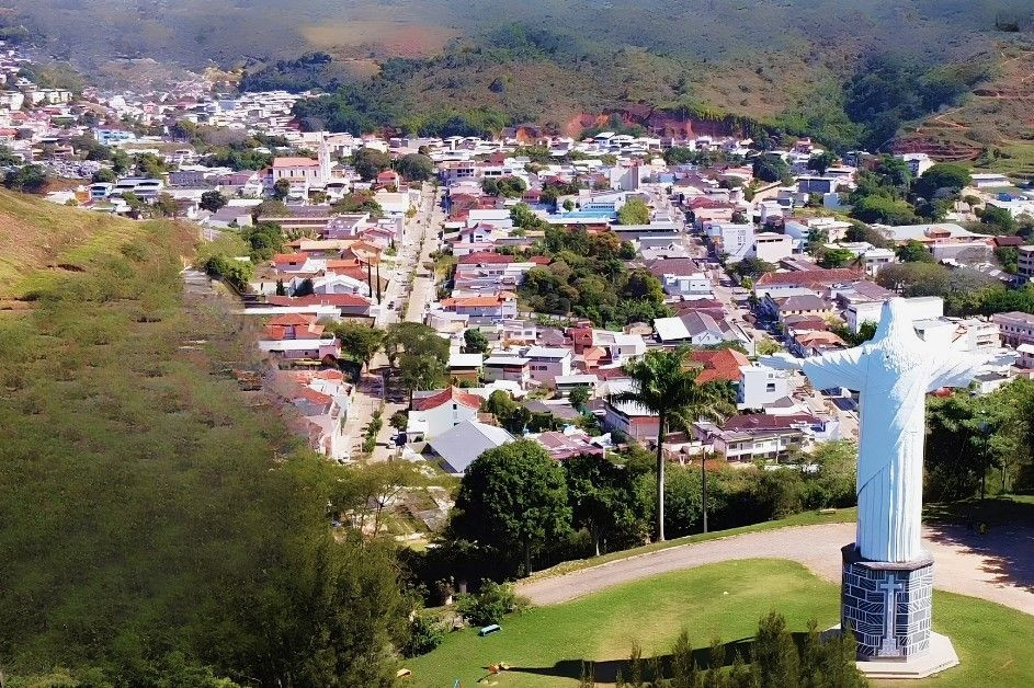 Vista aérea panorâmica da cidade de Guaçuí, na região do Caparaó, Espírito Santo. Em primeiro plano, no canto inferior direito, destaca-se o Monumento do Cristo Redentor, uma estátua branca sobre uma base de pedras escuras decorada com uma cruz. O monumento fica em uma área gramada no alto de um morro. Ao fundo, estende-se o vale com o mapeamento urbano do município, composto por casas, prédios de baixa altura e ruas arborizadas, cercados por montanhas de relevo ondulado e vegetação nativa sob a luz do dia.