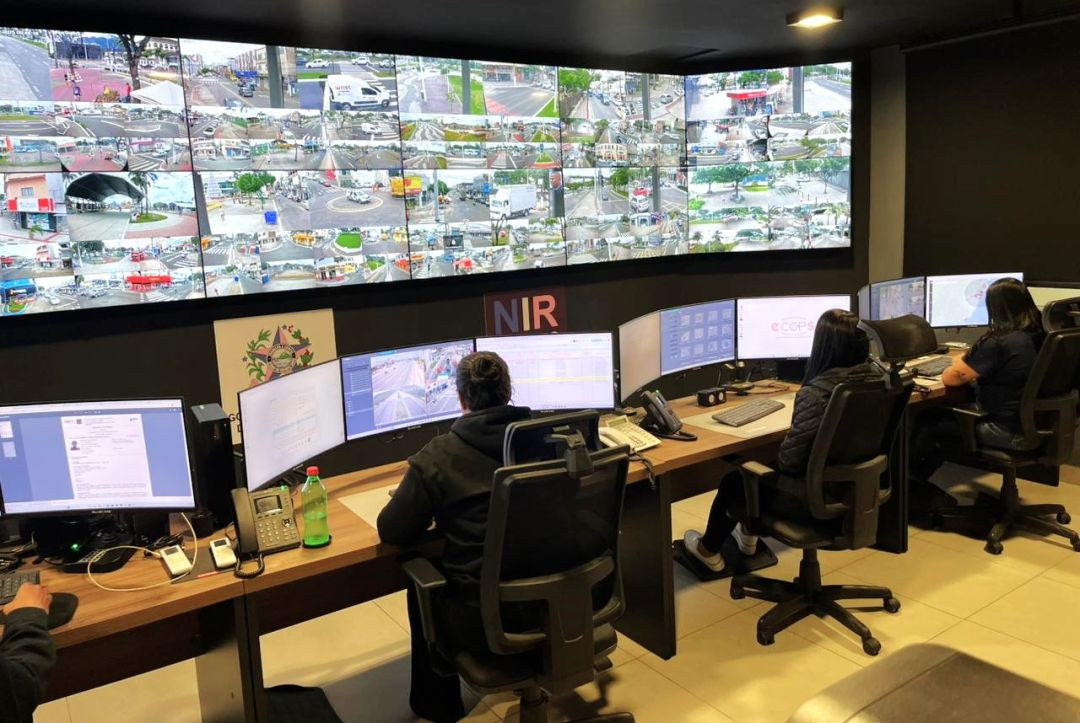 Sala de controle e monitoramento de segurança pública em ambiente com iluminação baixa. Ao fundo, uma grande parede convexa é composta por dezenas de telas menores que exibem imagens em tempo real de vias públicas, cruzamentos e movimentação urbana de veículos e pedestres. Na parte central, abaixo das telas principais, há a sigla 