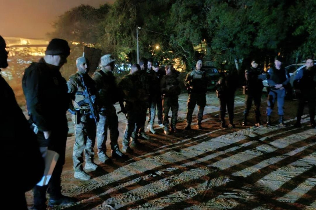 Alinhamento de policiais civis e militares em formação em um pátio de terra batida durante a madrugada, recebendo instruções antes do início da operação.