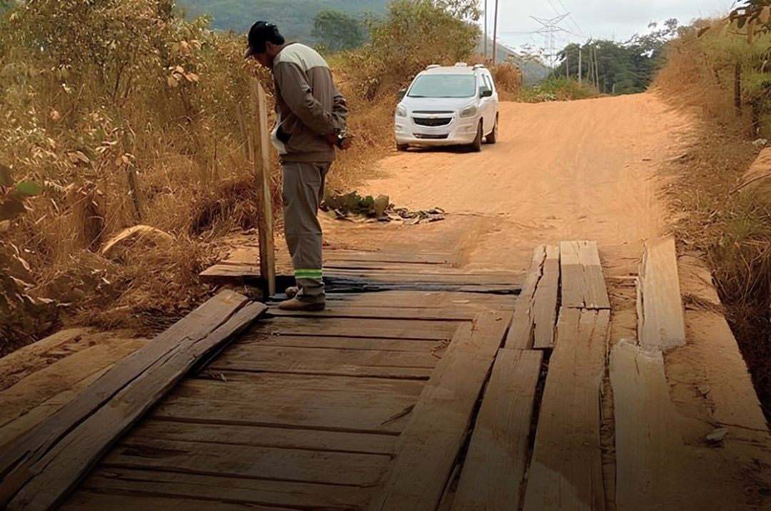 Homem usando boné e uniforme de trabalho observa e inspeciona as condições das tábuas de madeira que formam a pista de rolamento de uma ponte rural em estrada de terra. Ao fundo, um carro de passeio na cor branca aguarda na via cercada por vegetação seca e rasteira, em Terra Corrida, Iúna.