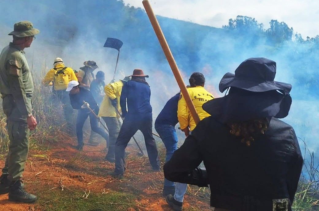Grupo de pessoas em uma encosta de terra e vegetação seca simulando o combate a um foco de incêndio com fumaça azulada ao fundo. Algumas vestem camisas amarelas com a inscrição IBAMA, outras usam chapéus de aba larga e operam ferramentas de manejo de solo e abafadores, sob a supervisão de um instrutor uniformizado à esquerda.