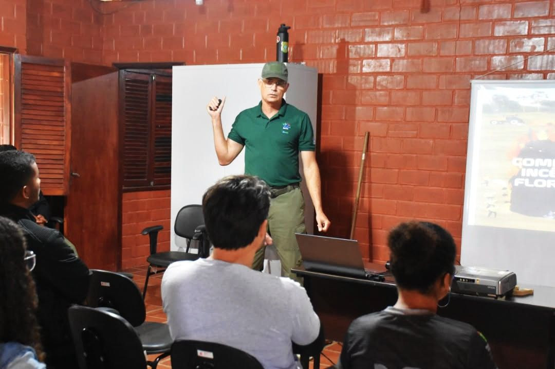 Homem com camisa polo verde e boné militar, identificado como instrutor, em pé diante de uma sala de aula de tijolos aparentes. Ele gesticula com uma das mãos enquanto ministra a palestra ao lado de uma projeção digital. Em primeiro plano, participantes assistem à instrução de costas.