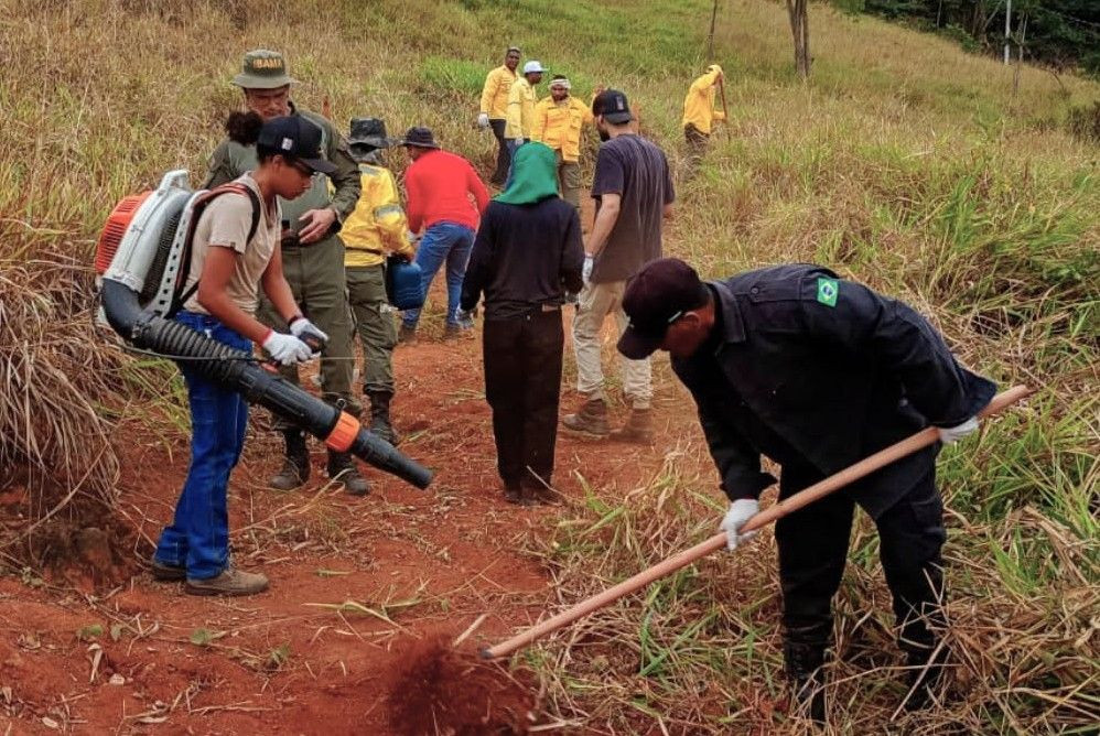 Atividade prática em terreno aberto com vegetação rasteira seca. No centro, voluntários utilizam ferramentas agrícolas como enxadas para limpar o solo e criar aceiros, enquanto uma mulher à esquerda carrega um soprador costal motorizado e recebe orientações de um técnico do IBAMA.