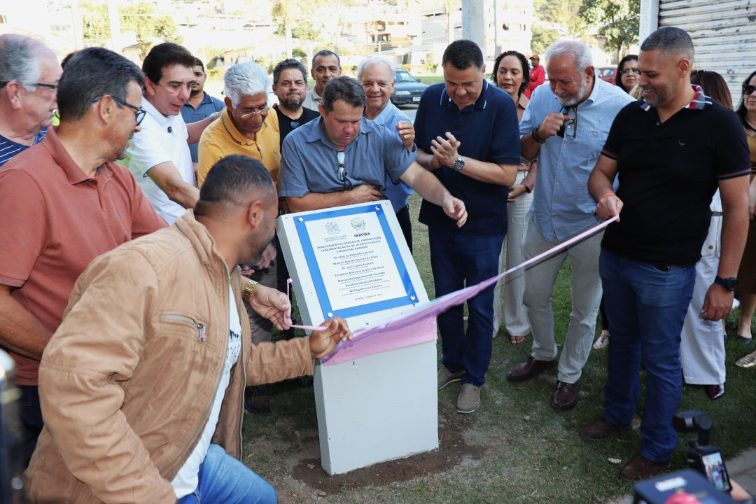 Grupo de homens e autoridades políticas reunidos ao ar livre em Ibatiba durante o descerramento de uma placa oficial de inauguração de obras nas avenidas Afonso Cláudio e Marechal Rondon. Um homem de jaqueta marrom agacha-se à esquerda puxando o tecido rosa que cobria a placa fixada em uma estrutura de concreto.