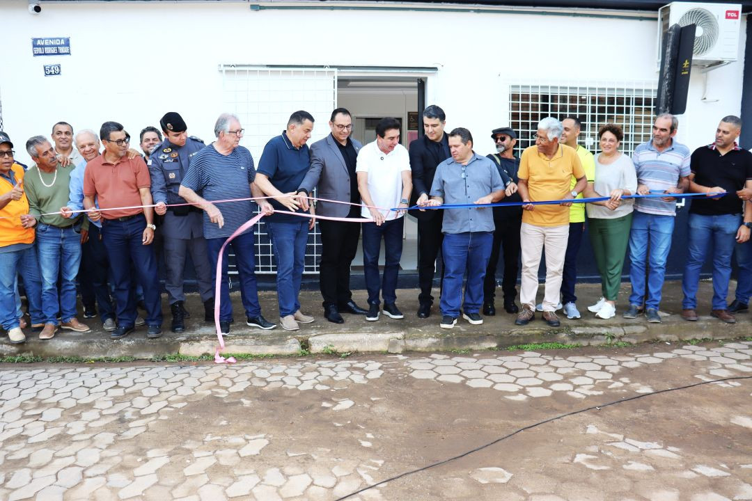 Autoridades municipais, estaduais e militares enfileiradas na calçada em frente à fachada de um prédio público com portas de grade branca. Juntos, eles seguram e cortam uma fita inaugural estendida nas cores rosa e azul, oficializando as entregas no município.