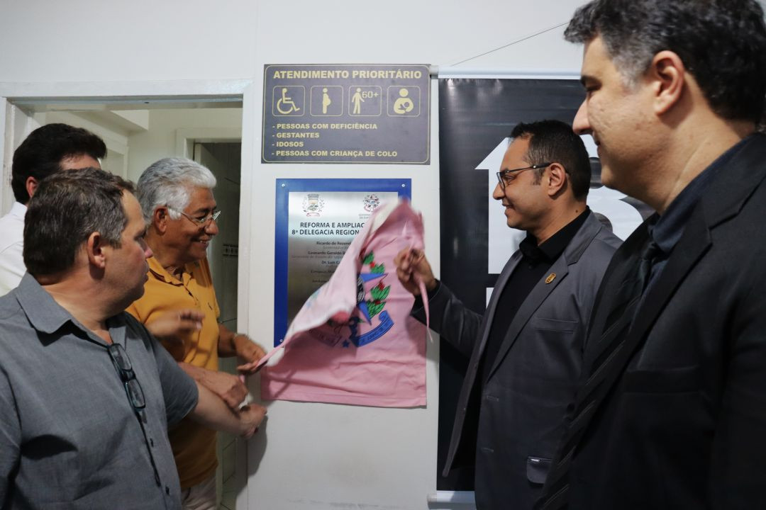 Registro em detalhe e de perfil do descerramento da placa de reforma e ampliação da 8ª Delegacia Regional de Polícia Civil no interior do prédio. Homens sorriem enquanto um deles puxa o pano rosa que cobria a placa metálica instalada na parede branca, abaixo de uma sinalização de atendimento prioritário.