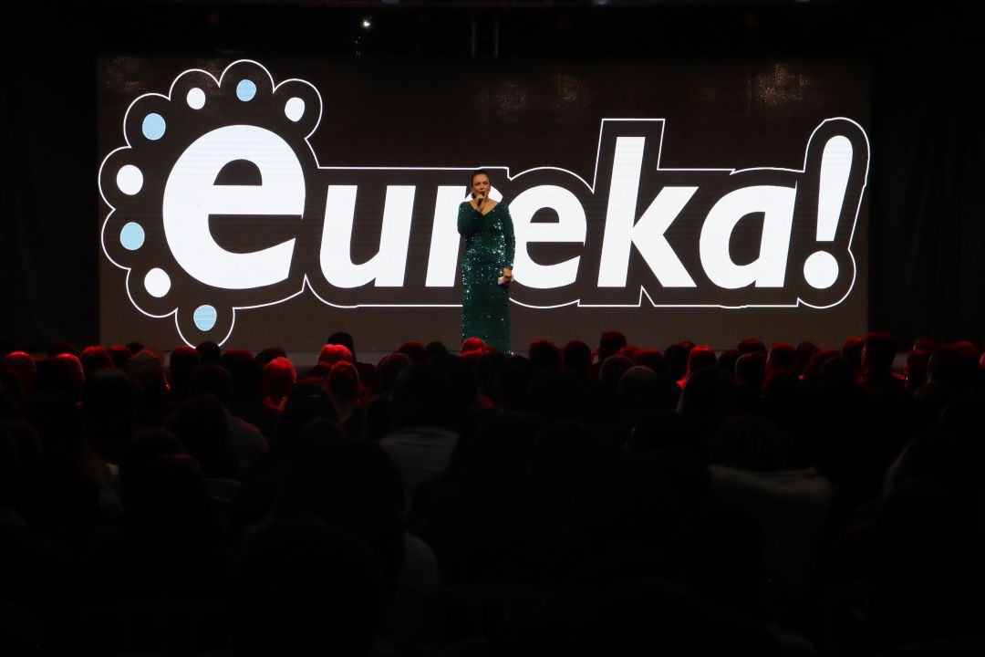 Mulher com vestido longo verde brilhante fala ao microfone no centro do palco. Ao fundo, um grande painel de LED exibe o logotipo estilizado do evento escrito 