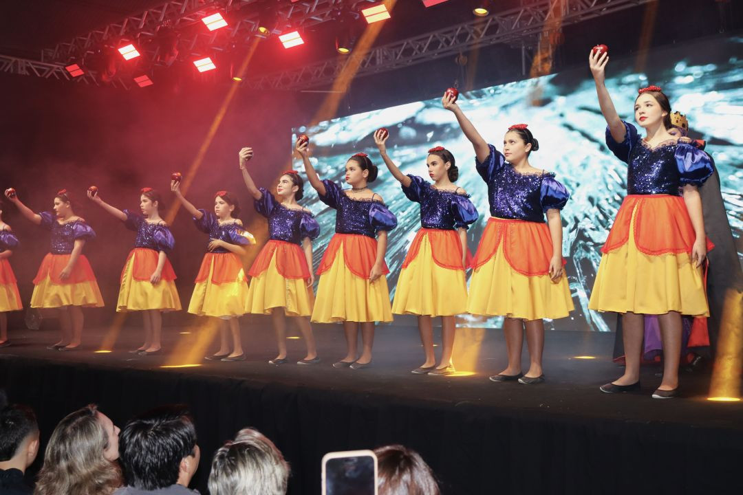 Uma fileira de alunas perfiladas no palco, vestidas com fantasias inspiradas na Branca de Neve (corpete azul brilhante com mangas bufantes e saia amarela e vermelha). Cada uma segura uma maçã vermelha no alto com a mão esquerda. O palco está iluminado por fortes refletores de luz vermelha e amarela, com a plateia visível em primeiro plano abaixo.