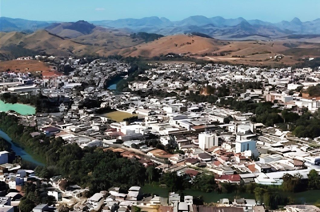 Fotografia aérea em plano aberto da cidade de Bom Jesus do Norte. Em destaque, a área urbana com grande concentração de residências e edifícios de tons claros. Um rio de águas esverdeadas serpenteia a cidade, margeado por árvores e áreas verdes. Ao fundo, erguem-se morros de terra batida e montanhas em tons de azul e marrom sob um céu claro, caracterizando o relevo da região do Caparaó.